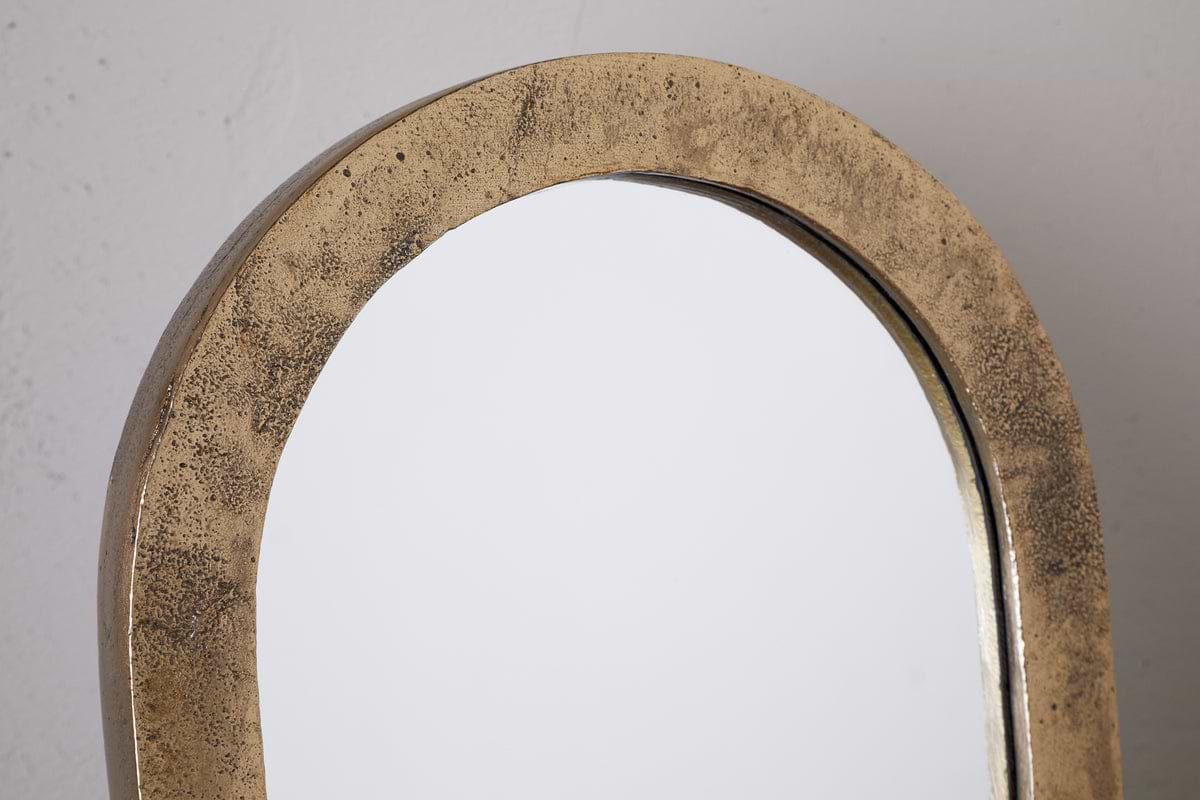 A close-up of the Cayce Cast Aluminum Vanity Mirror by nkuku, featuring an arched top and textured antique brass frame, set against a plain light background.
