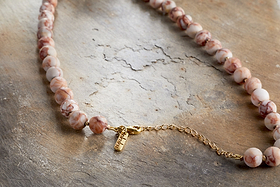 The Cerita Necklace - Rhodochrosite by nkuku features marbled pink and white semi-precious stones and a gold chain extender, displayed on an earthy textured surface—a versatile accessory for any occasion.