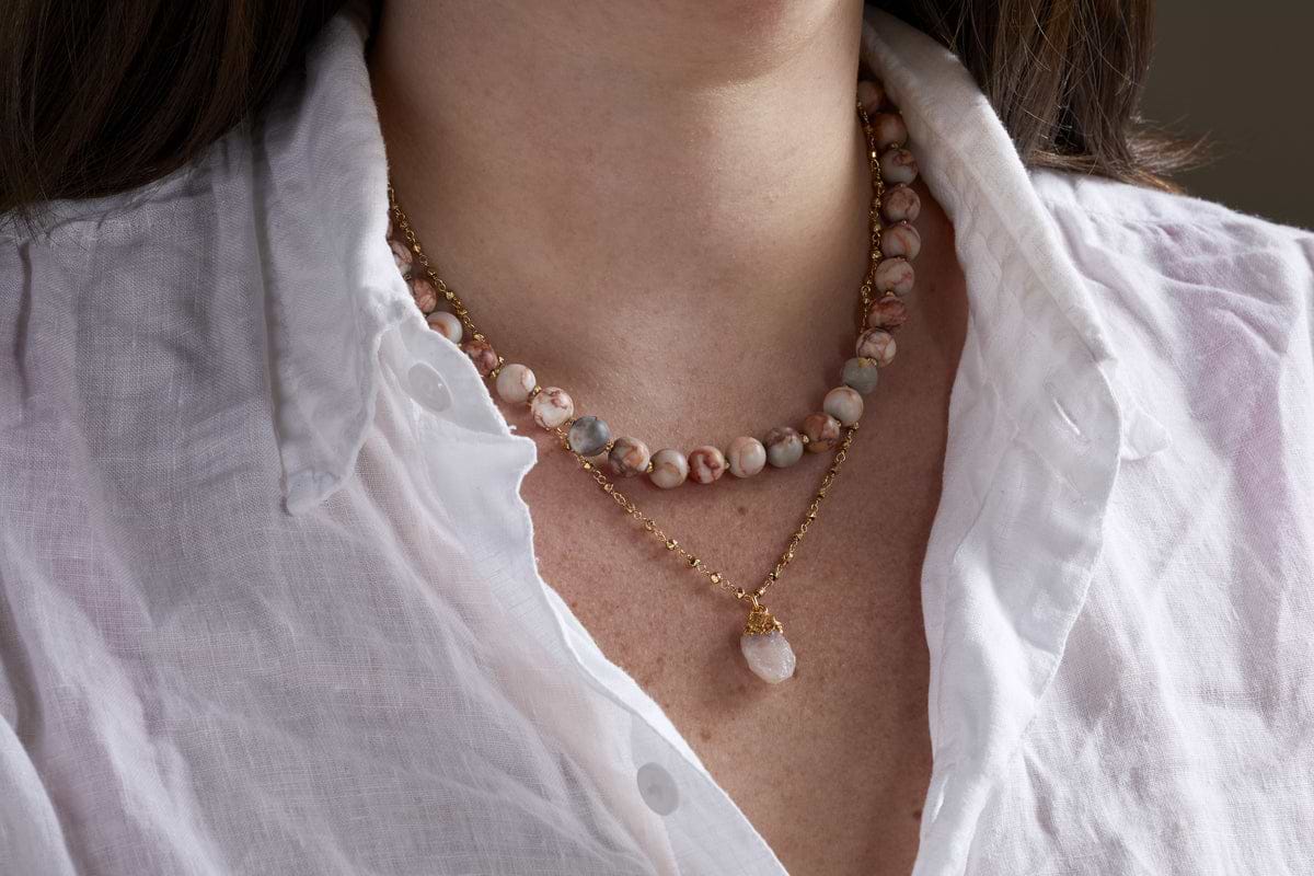 A woman in a white button-up shirt wears two layered necklaces, including nkukus Cerita Necklace - Rhodochrosite: a gold chain with a pink rhodochrosite pendant, paired with semi-precious stone beads for an effortlessly versatile look.