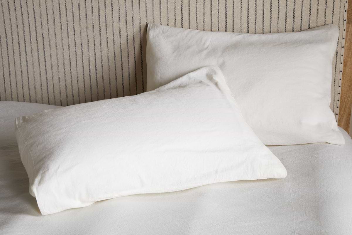 Two Cela Hemp & Cotton Housewife Pillowcases in natural by nkuku rest on a neatly made bed with a light, vertically striped headboard, creating a clean and minimalistic bedroom style.