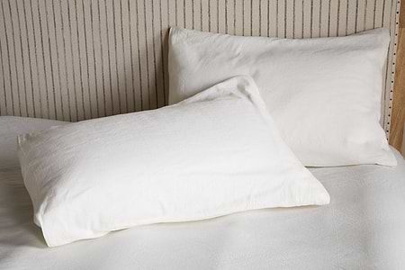 Two Cela Hemp & Cotton Housewife Pillowcases in natural by nkuku rest on a neatly made bed with a light, vertically striped headboard, creating a clean and minimalistic bedroom style.