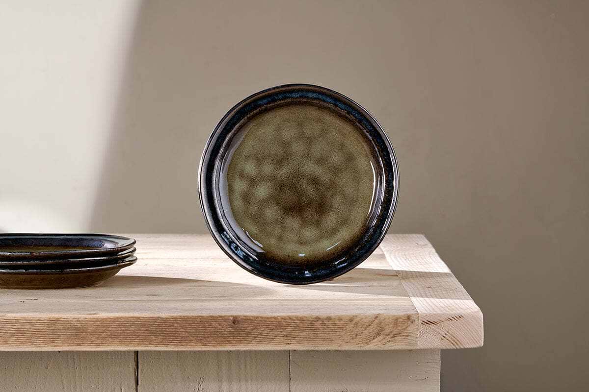 The Cali Side Plate - Earth Blue by nkuku, with a mottled blue center and glossy dark rim, stands upright on a light wooden table. Two matching cafe-style plates are stacked on the left. The beige wall behind casts a shadow to the left.
