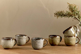 Five nkuku Cam Coffee Mugs - Mocha (Set of 2) with reactive glaze are arranged on a wooden surface; three stand alone, two are stacked. A glass vase with dried greenery is set in the background against a beige wall.