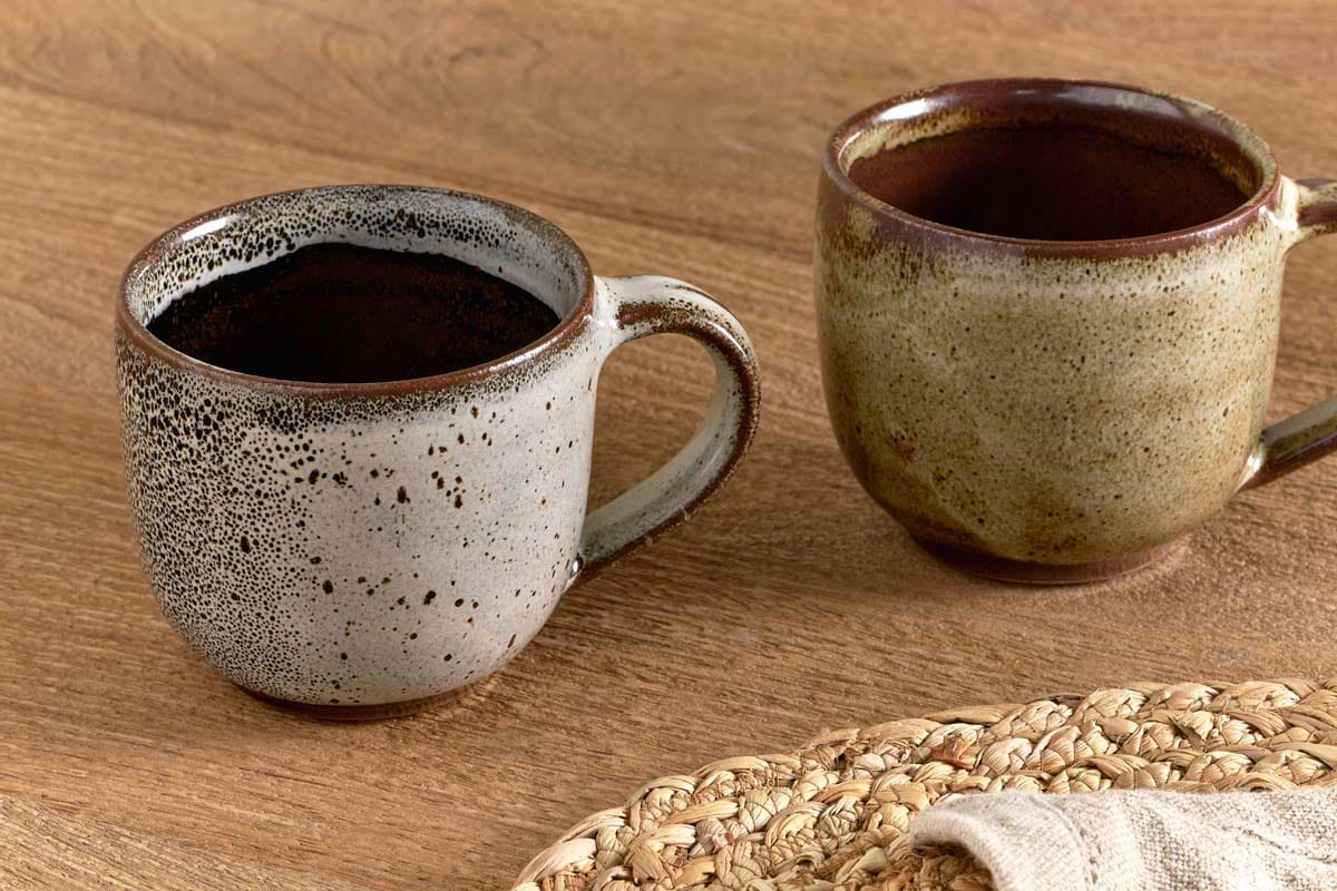 The nkuku Cam Coffee Mug - Mocha (Set of 2) features two rustic mugs, one gray with black speckles and one olive green glaze, on a wooden table with coffee, a woven placemat, and napkin for a cozy setting.