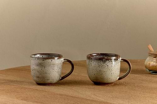 Two Cam Coffee Mugs - Mocha by nkuku (set of 2) with a speckled reactive glaze rest on a wooden table. A glass jar with a wooden lid appears on the right, all set against a neutral beige wall backdrop.
