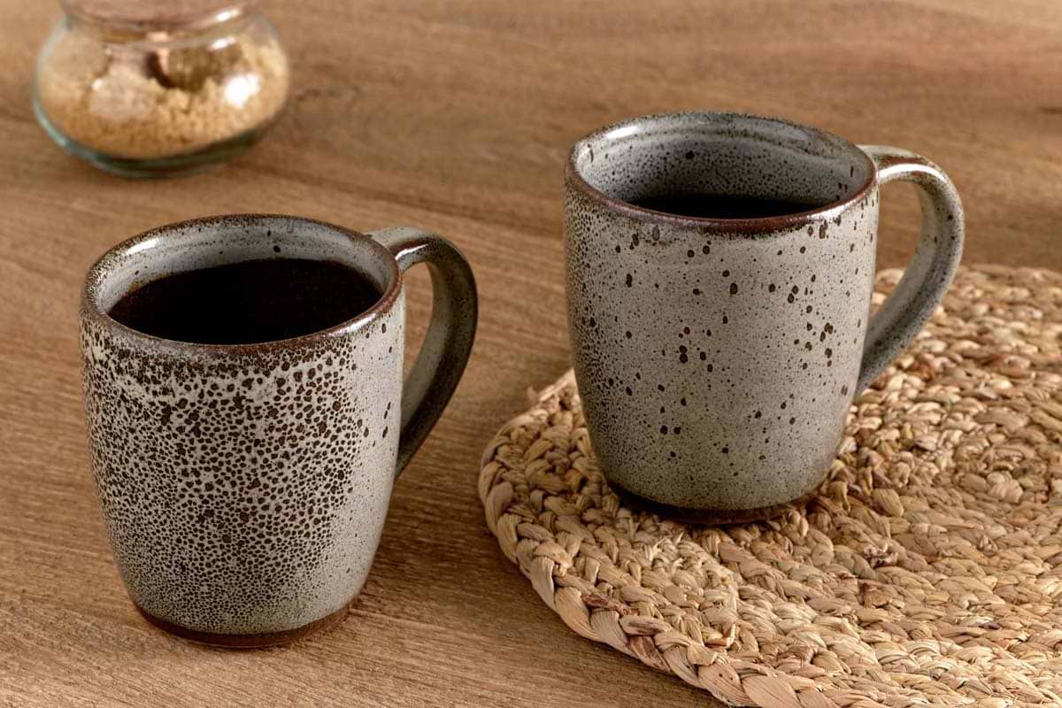 Two nkuku Cam Large Mugs - Mocha (Set of 2) filled with dark coffee sit on a wooden table, one on a woven placemat. A brown sugar jar in the background highlights the charm of these speckled grey reactive glaze mugs.