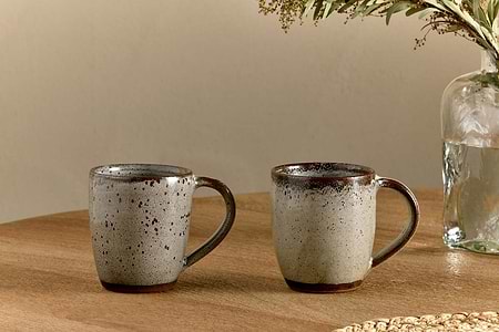 Two nkuku Cam Large Mug - Mocha (Set of 2) with brown rims and speckled gray glaze rest side by side on a wooden table. A glass vase with water and green foliage is partially visible in the background.