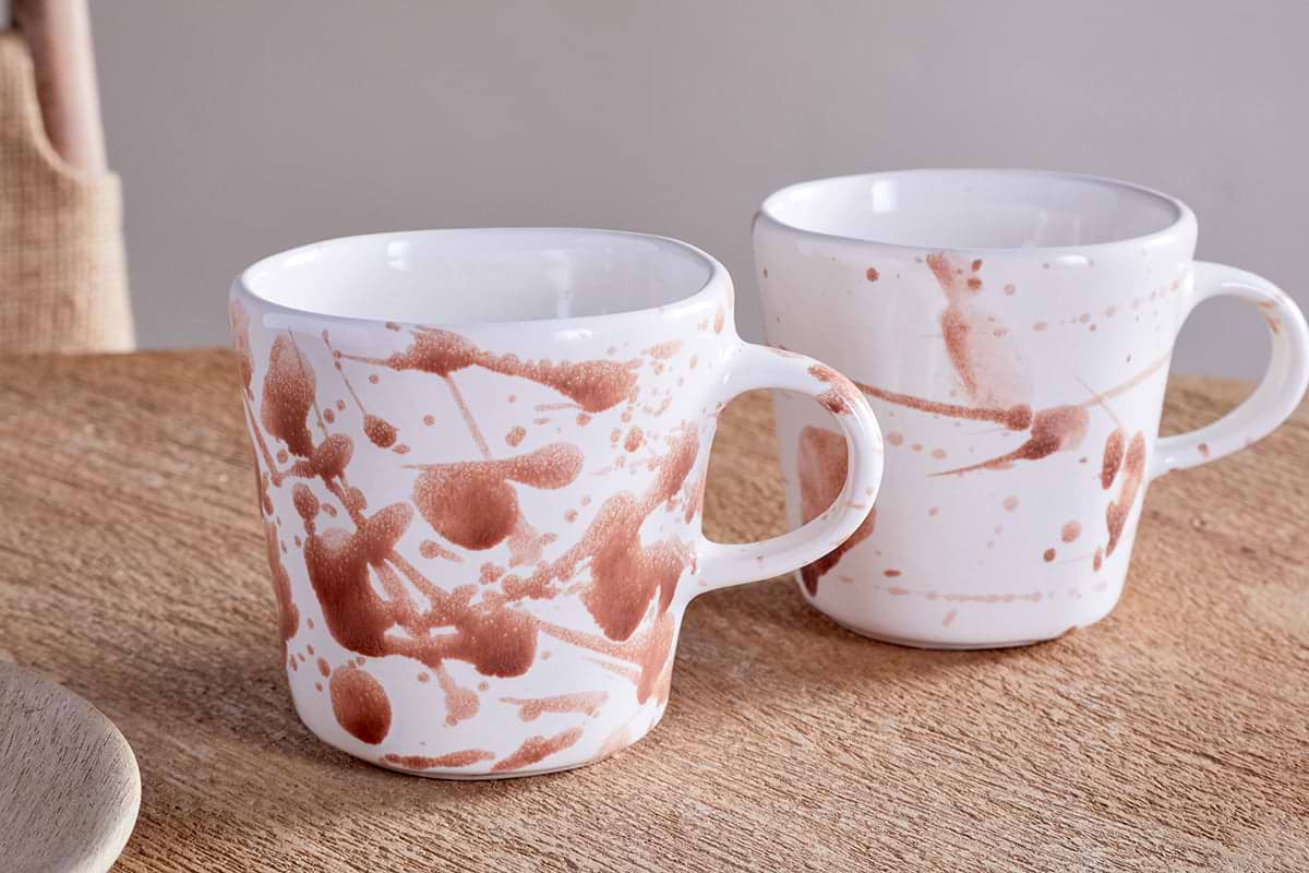 The nkuku Canela Mug set in Dusty Terracotta features two ceramic mugs with brown abstract splatter designs, offering a cozy, minimalist touch to your space. Perfect for enjoying beverages together or as a stylish gift.