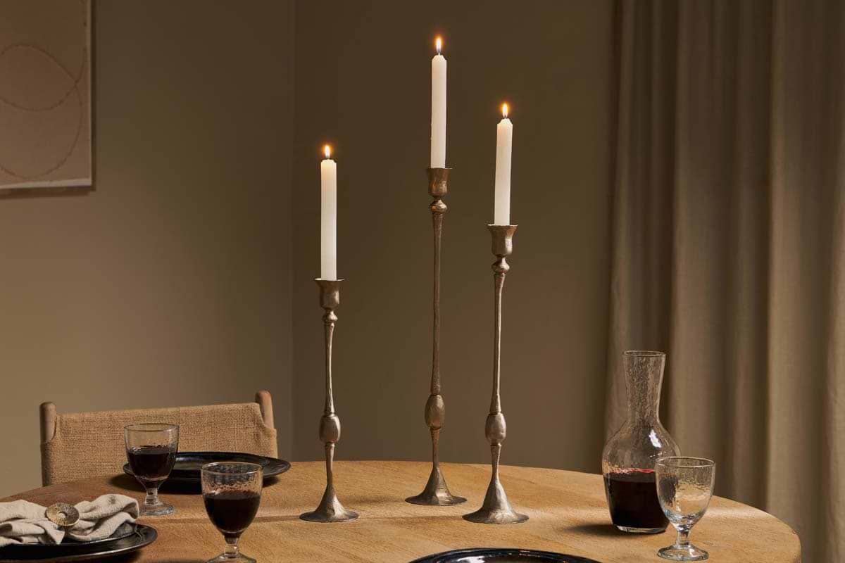 Three lit white candles in nkuku's Chaipat Candlesticks in antique brass are centered on a round wooden table, which also features two red wine glasses, glassware, a clear carafe, black plates, and a beige napkin against warmly-lit neutral walls.