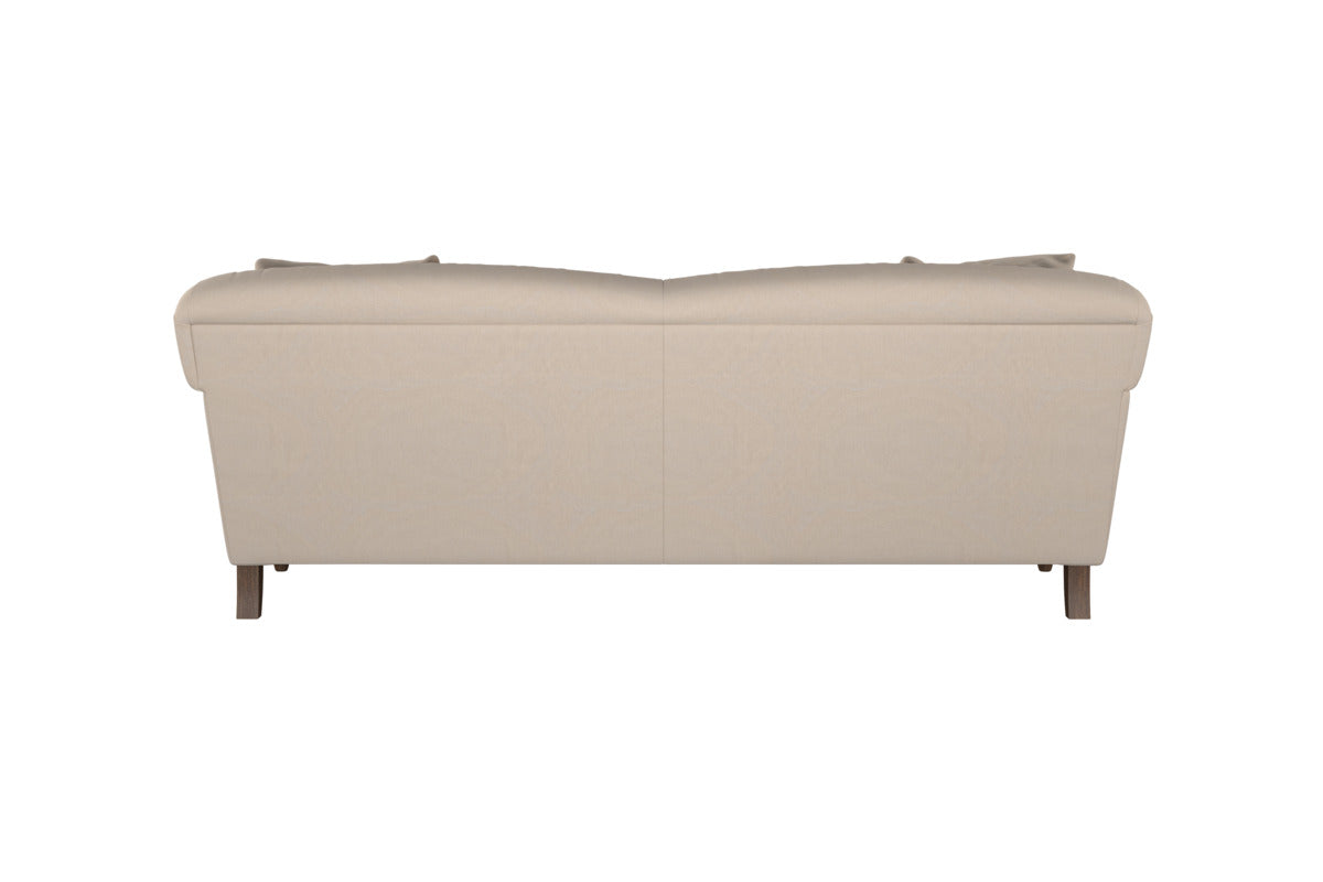 The Nkuku Deni Large Corner Sofa in Brera Linen Smoke is shown from the back, showcasing its smooth linen upholstery in a beige tone. It features gently rounded armrests and short wooden legs, while the plush backrest cushions and simple, modern lines reflect superior British quality. The sofa is set against a white background.