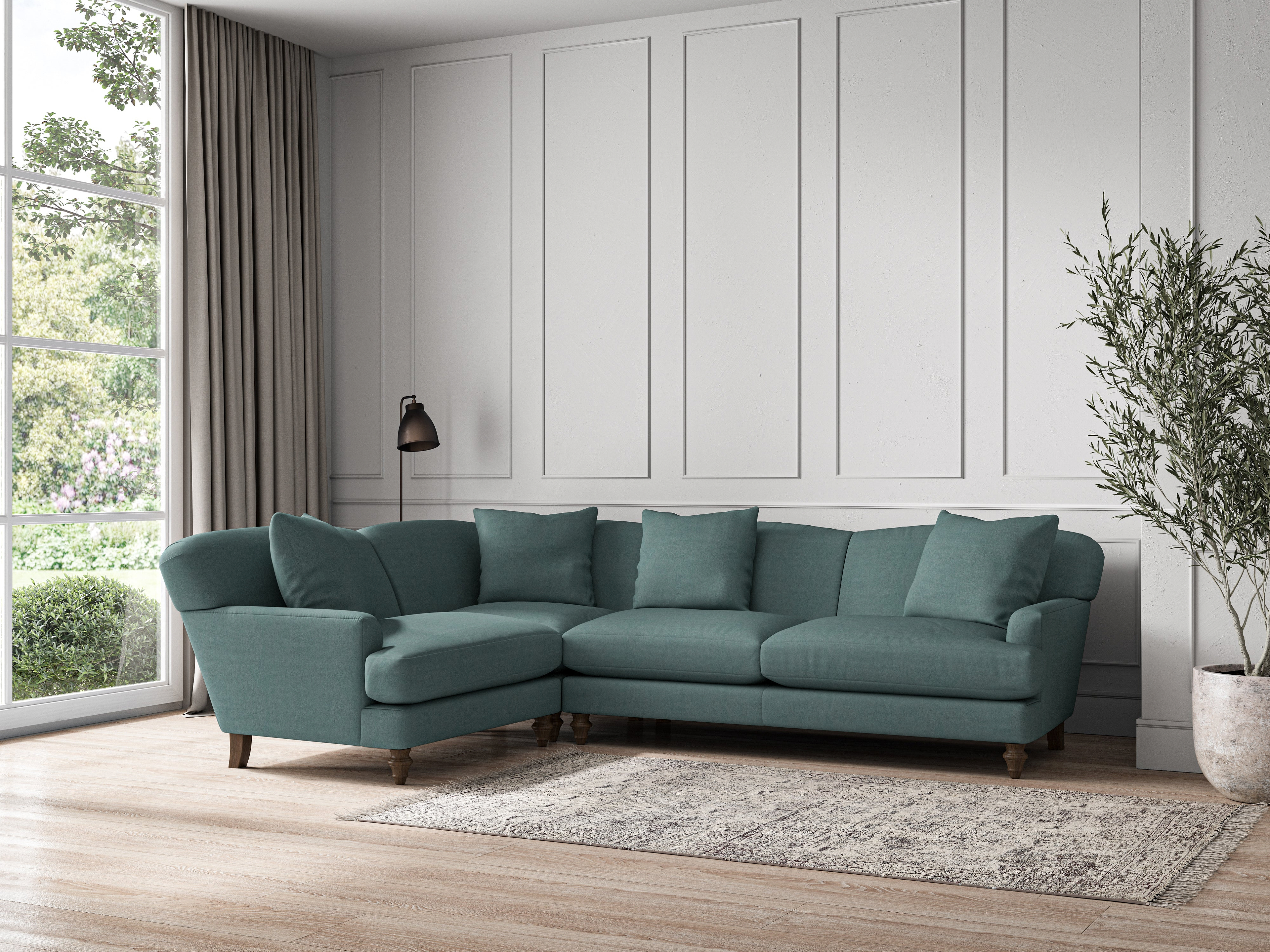 A modern living room features the nkuku Deni Large Left Hand Corner Sofa in recycled cotton Airforce on a light rug, set against white paneled walls. Beige curtains frame a large window, with a black floor lamp and tall green plant accenting the space.