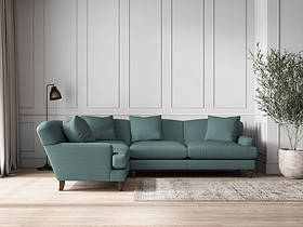 A modern living room showcases the nkuku Deni Large Left Hand Corner Sofa in Recycled Cotton Airforce, featuring five cushions against a white paneled wall. To the right is a potted olive tree; to the left, a black floor lamp and a light rug on wood floors.