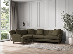 A modern living room featuring the nkuku Deni Large Left Hand Corner Sofa in Recycled Cotton Fatigue, four matching cushions, a textured rug, floor lamp, tall curtains, wood flooring, indoor plant, white paneled walls, and window views of greenery.