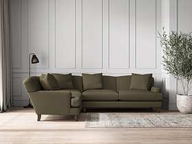 A modern living room features the nkuku Deni Large Left Hand Corner Sofa in recycled cotton fatigue, four matching cushions, a gray area rug, floor lamp, potted plant, and wood flooring. White paneling lines the back wall; light enters from curtained windows.