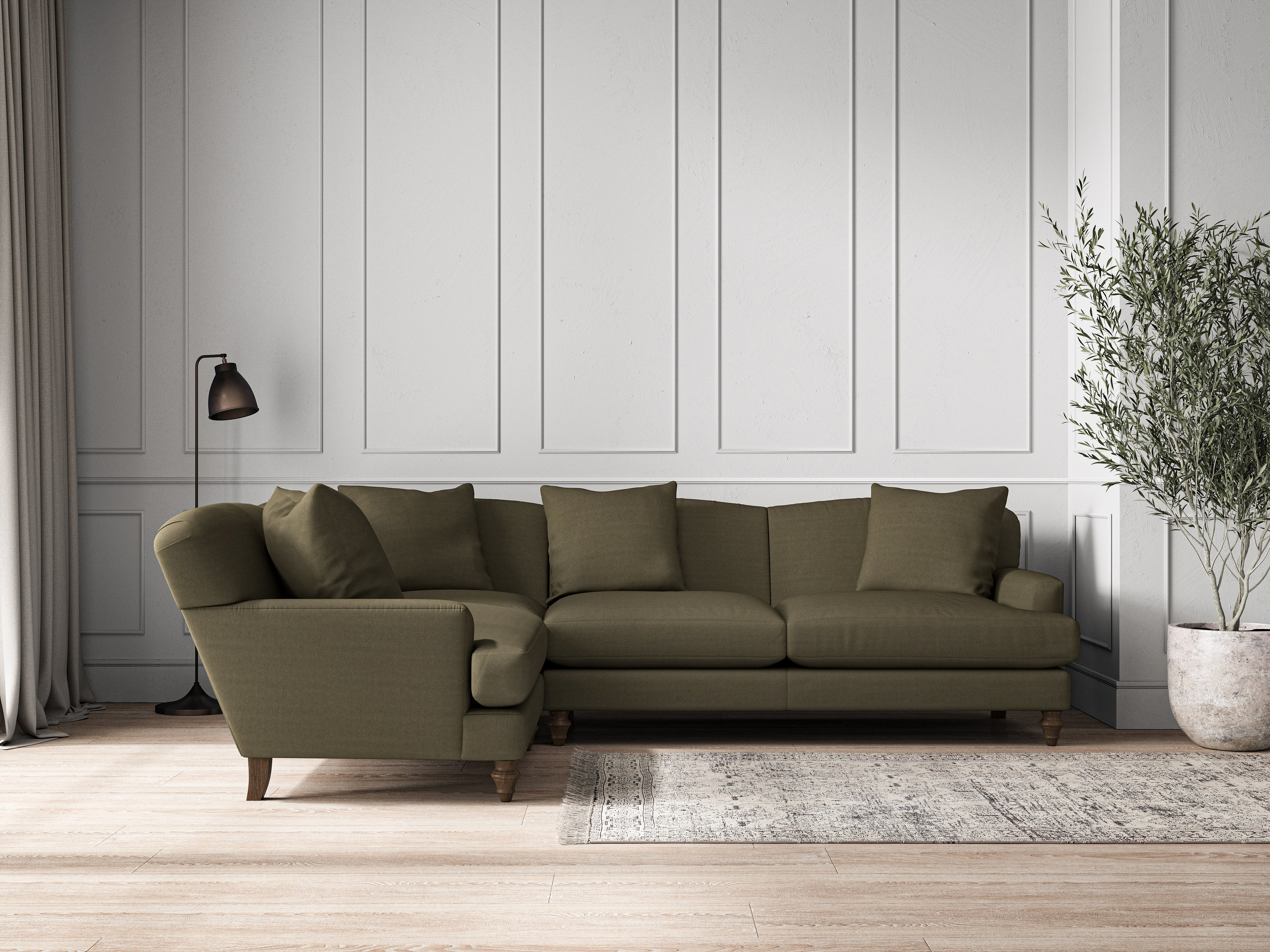 A modern living room features the nkuku Deni Large Left Hand Corner Sofa in recycled cotton fatigue, four matching cushions, a gray area rug, floor lamp, potted plant, and wood flooring. White paneling lines the back wall; light enters from curtained windows.