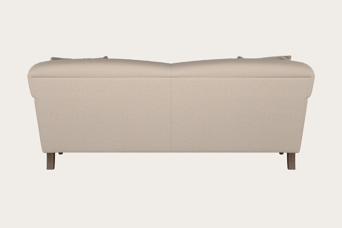 A rear view of the Nkuku Deni Large Sofa in Recycled Cotton Natural showcases its smooth, beige upholstery, with two seat back cushions subtly visible above. The modern design is complemented by four wooden legs and exemplifies British quality furniture, set against a plain off-white background.