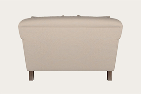 A rear view of the Nkuku Deni Love Seat - Recycled Cotton Natural reveals its smooth, padded beige upholstery and gently rounded top edges. The slightly tapered sides and two visible wooden legs highlight the craftsmanship typical of handcrafted hardwood furniture. The background is plain and light-colored.