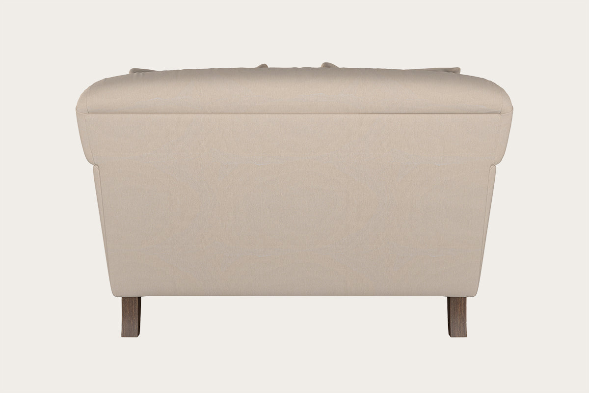 A rear view of the Nkuku Deni Love Seat - Recycled Cotton Natural reveals its smooth, padded beige upholstery and gently rounded top edges. The slightly tapered sides and two visible wooden legs highlight the craftsmanship typical of handcrafted hardwood furniture. The background is plain and light-colored.
