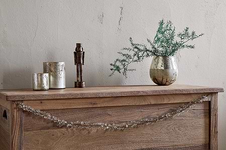 A rustic wooden chest features the nkuku Danvi Glass Drop Garland in brass and clear draped across the front. On top sit two metallic candle holders, a bronze figurine, and a silver vase with green foliage, set against a textured beige wall.