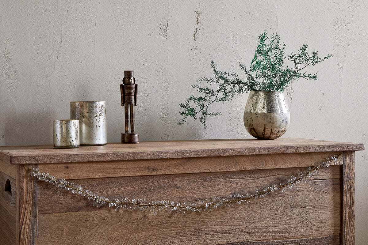 A rustic wooden chest features the nkuku Danvi Glass Drop Garland in brass and clear draped across the front. On top sit two metallic candle holders, a bronze figurine, and a silver vase with green foliage, set against a textured beige wall.