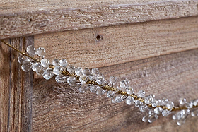 A close-up of a wooden surface decorated with the nkuku Danvi Glass Drop Garland in Brass & Clear, featuring twisted gold wire and crystal-like glass drops draped diagonally for a festive look.