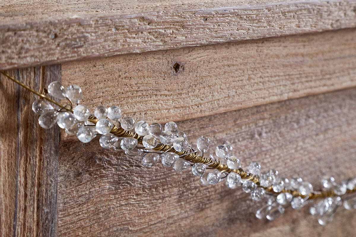 A close-up of a wooden surface decorated with the nkuku Danvi Glass Drop Garland in Brass & Clear, featuring twisted gold wire and crystal-like glass drops draped diagonally for a festive look.