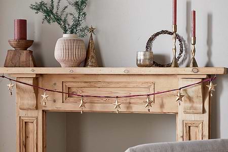 A rustic wooden mantel is decorated with nkuku’s Didi Star Garland in antique brass, two red candles, a pink-striped vase of greenery, a metallic cone tree, a round twig wreath, and a small metallic cup against a light beige wall.