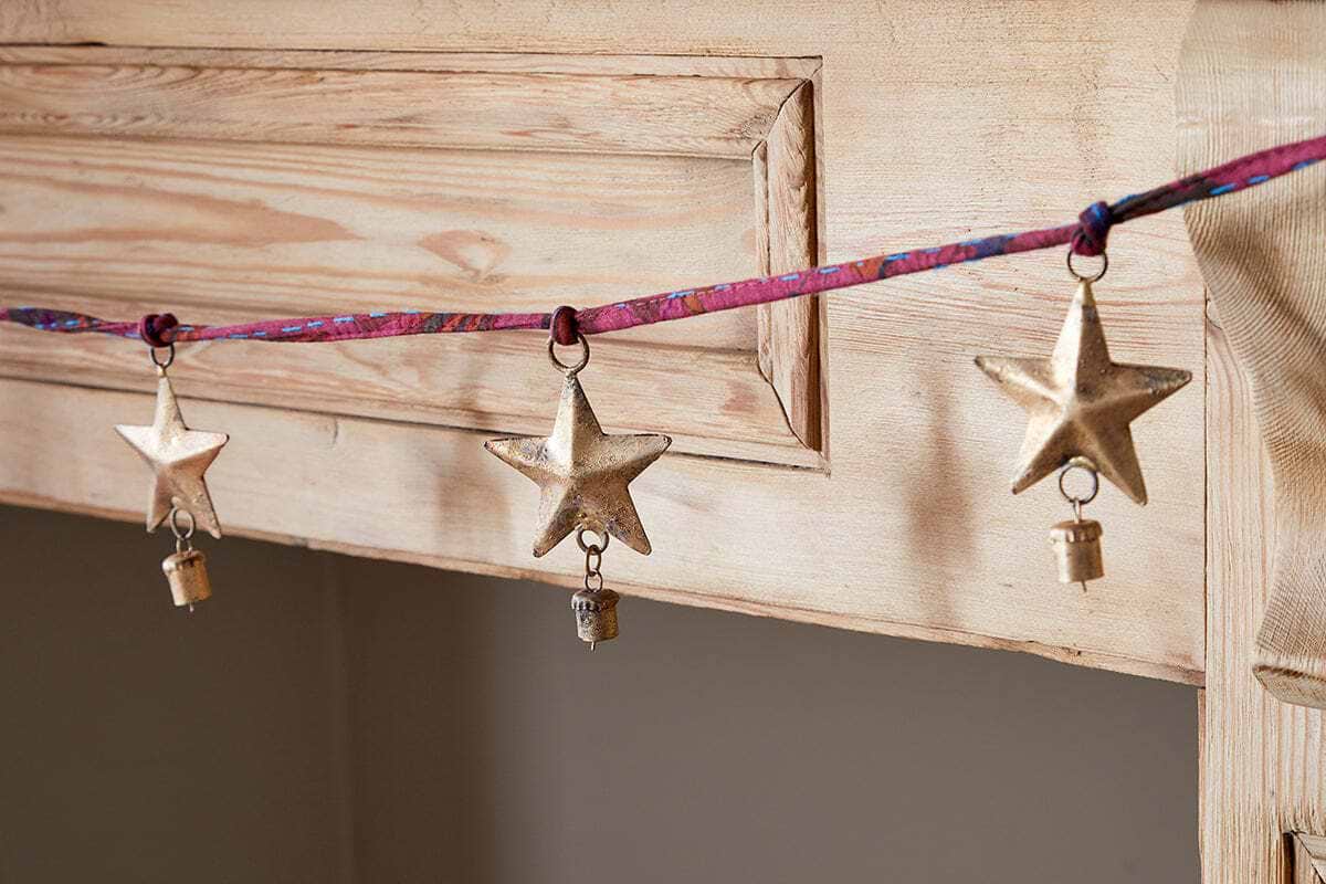 The nkuku Didi Star Garland features three handcrafted antique brass stars, each with a small key, hanging from a colorful ribbon and displayed on natural wood for a festive, artisanal touch.