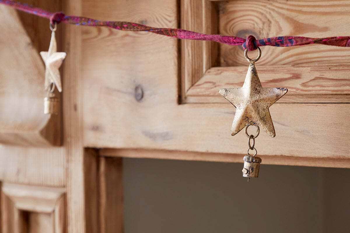 A close-up of the nkuku Didi Star Garland - Antique Brass, featuring antique brass stars and small bells on a twisted pink and red cord, draped over a rustic wooden cabinet with a softly blurred background.