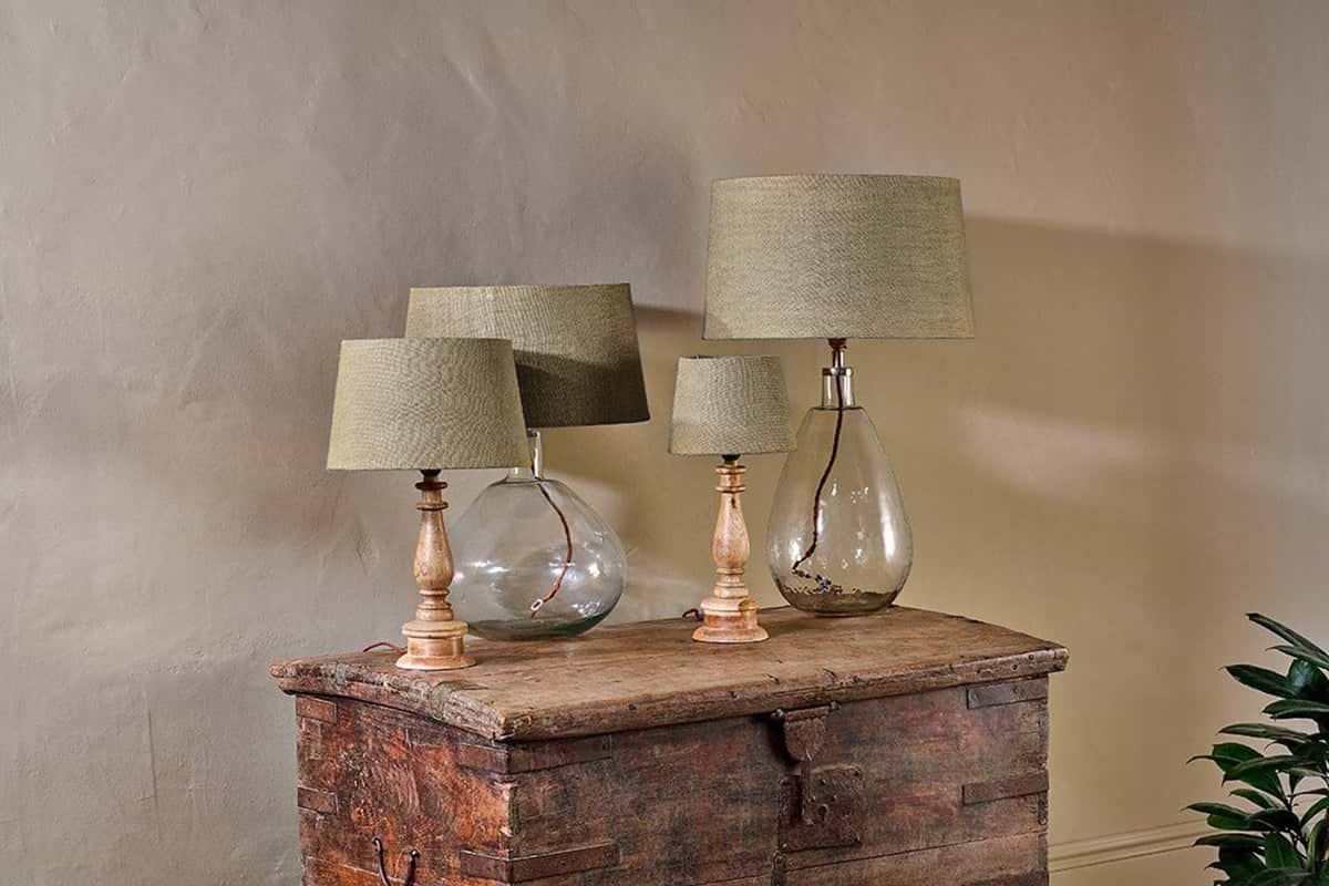 Three nkuku Dia Jute Lampshade - Natural table lamps with glass and wood bases sit on a rustic wooden chest against a neutral wall, casting a warm ambient glow. A leafy green plant is partially visible in the lower right corner.