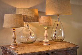 Three nkuku Dia Jute Lampshade table lamps with natural jute shades sit on a rustic chest. Two have clear, textured glass bases; one features a carved wooden base. A green plant appears at the bottom right.
