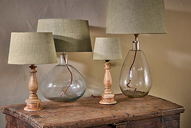 Four nkuku Dia Jute Lampshade - Natural table lamps sit on a rustic wooden chest. Two feature clear glass bases, two have turned wood with a textured finish. Their ambient glow creates a cozy atmosphere against the beige wall.