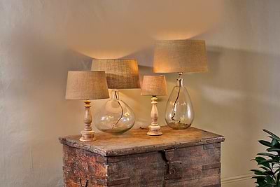 Four nkuku Dia Jute Lampshade - Natural table lamps sit on a rustic wooden surface. Two have clear glass bases (round and teardrop); two feature light wood bases. Visible cords add charm, making them ideal for ambient lighting.