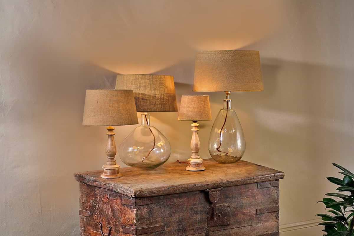 Four nkuku Dia Jute Lampshade - Natural table lamps sit on a rustic wooden surface. Two have clear glass bases (round and teardrop); two feature light wood bases. Visible cords add charm, making them ideal for ambient lighting.