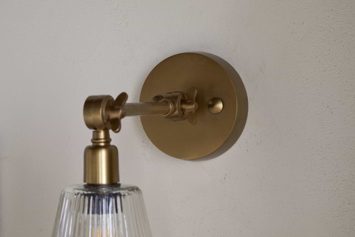 The nkuku Davika Ribbed Glass Task Wall Lamp in clear and antique brass features a round backplate, adjustable arm, and clear ribbed glass shade, shown close-up on a light textured wall.