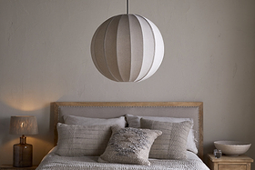 A neatly made bed with neutral bedding and textured pillows sits against a pale wall. Above, the nkuku Dharini Linen Lampshade in Natural hangs as the bedroom light. A wooden nightstand on the right holds a bowl; the left one has a lamp.