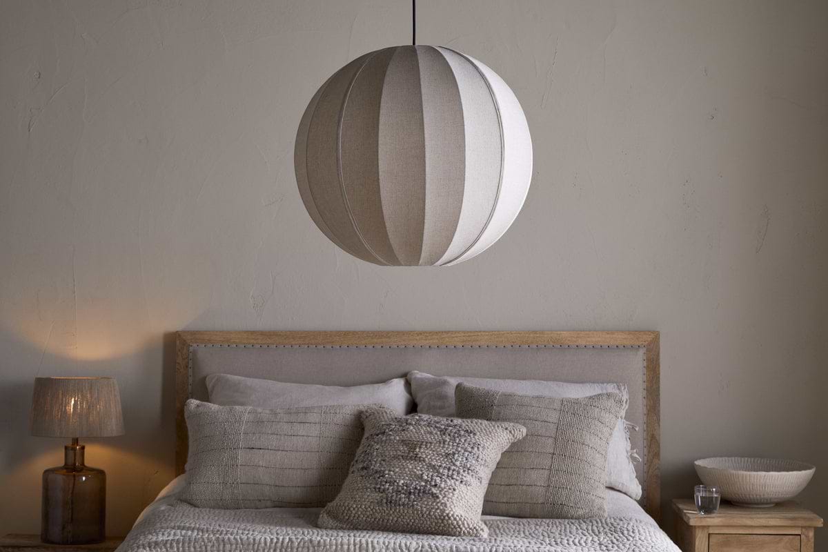 A neatly made bed with neutral bedding and textured pillows sits against a pale wall. Above, the nkuku Dharini Linen Lampshade in Natural hangs as the bedroom light. A wooden nightstand on the right holds a bowl; the left one has a lamp.