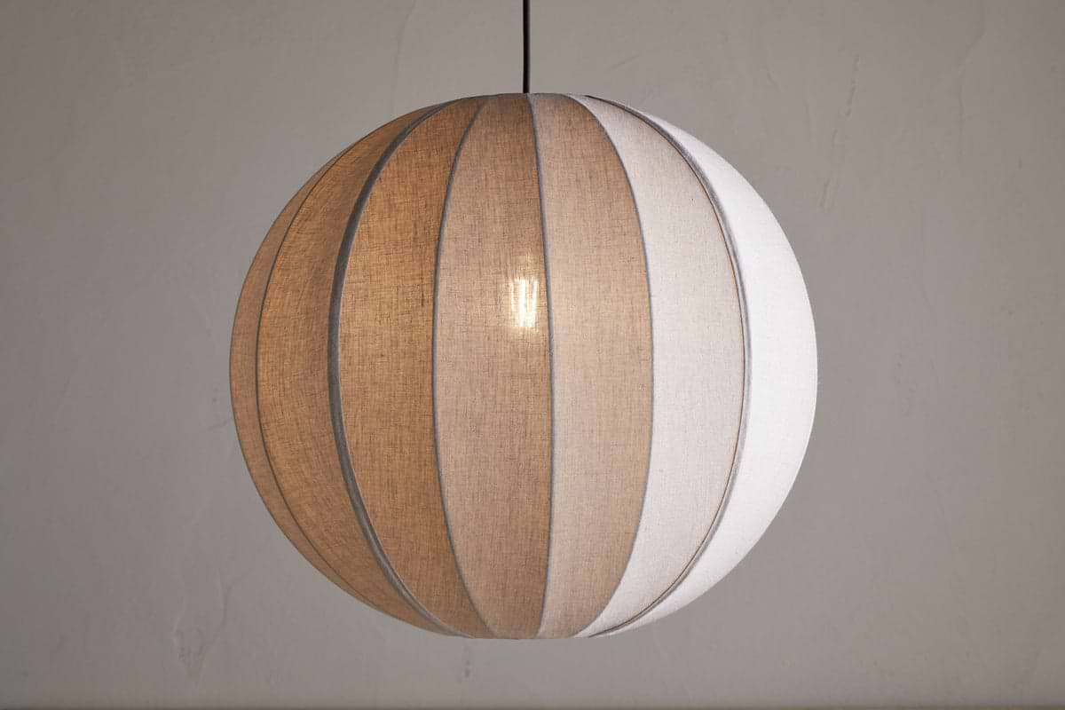 The Dharini Linen Lampshade - Natural by nkuku features a spherical linen shade with beige and white panels, suspended from a black cord against a light gray wall, softly illuminating your bedroom with its warm, textured glow.