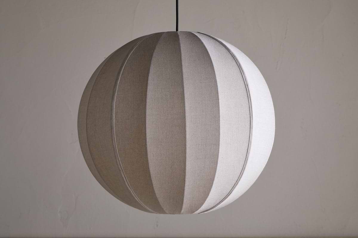 The nkuku Dharini Linen Lampshade - Natural features a round silhouette with vertical, alternating light and dark linen panels, suspended from a black cord. Its woven texture suits any elegant setting, ideal for bedroom ambiance.