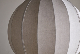 The Dharini Linen Lampshade - Natural by nkuku features a spherical, beige, textured linen design with vertical seams for a segmented look. Its simple backdrop highlights the lampshade’s elegant structure—perfect for bedroom lighting.