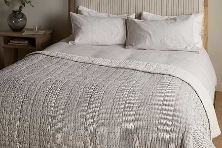 A neatly made bed with light beige and white linens, two pillows, and the nkuku Dipti Cotton & Linen Block Print Bed Quilt in charcoal. A wooden headboard and a small nightstand with a glass vase and water sit beside the bed.