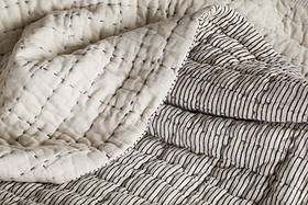 A close-up of the nkuku Dipti Cotton & Linen Block Print Bed Quilt - Charcoal shows two patterns: scattered horizontal stitches on cream fabric and neat vertical charcoal stripes on a light background.