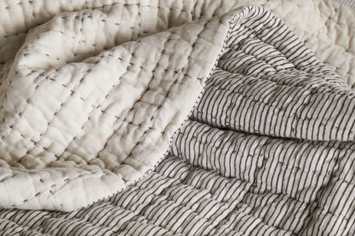 A close-up of the nkuku Dipti Cotton & Linen Block Print Bed Quilt - Charcoal shows two patterns: scattered horizontal stitches on cream fabric and neat vertical charcoal stripes on a light background.