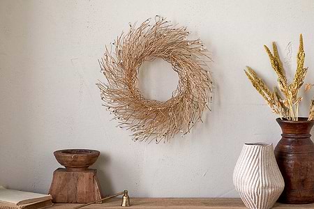 A minimalist scene showcases nkuku’s Danvi Glass Drop Wreath – Brass & Clear, hanging on a neutral wall above a rustic wood surface with a wooden bowl, striped ceramic vase, brown vase with dried grasses, brass bell, and closed book.