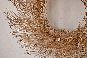 The Danvi Glass Drop Wreath by nkuku features intertwined brass wires shaped into a circle, with clear glass beads resembling dewdrops. Shown here against a plain, light beige wall.