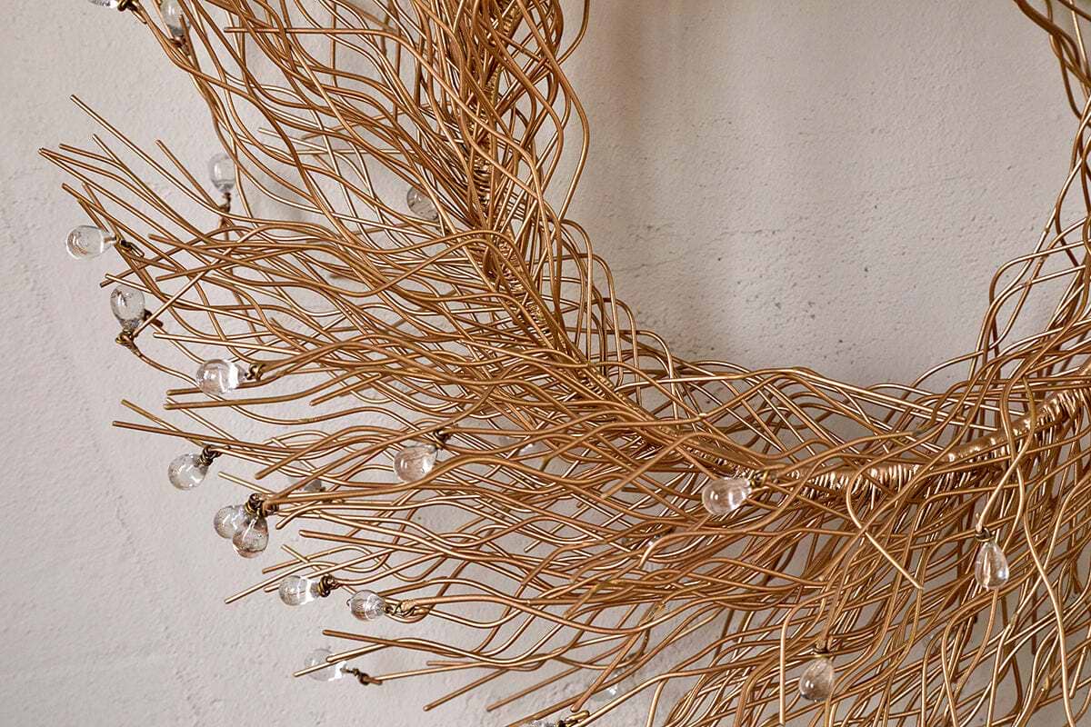 The Danvi Glass Drop Wreath by nkuku features intertwined brass wires shaped into a circle, with clear glass beads resembling dewdrops. Shown here against a plain, light beige wall.