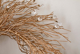 Close-up of the Danvi Glass Drop Wreath by nkuku, featuring intertwined brass wires with clear glass beads, creating a delicate, organic form against a light-colored wall.