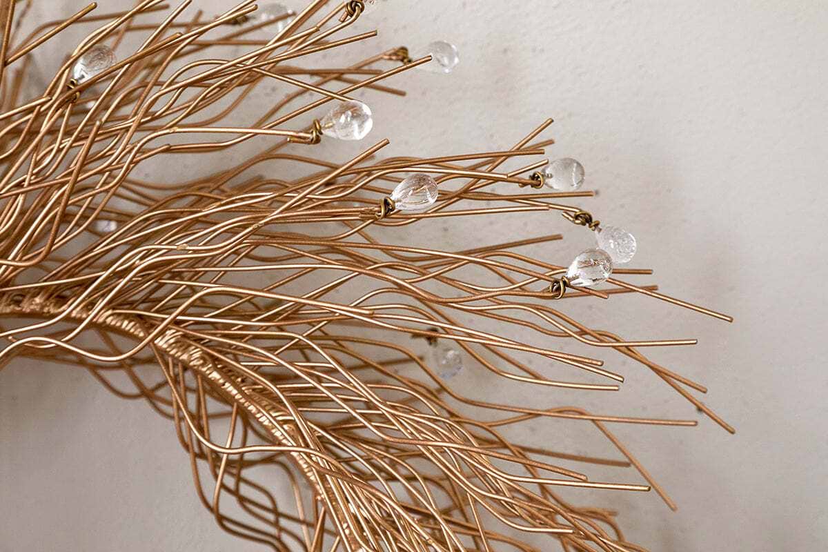 Close-up of the Danvi Glass Drop Wreath by nkuku, featuring intertwined brass wires with clear glass beads, creating a delicate, organic form against a light-colored wall.