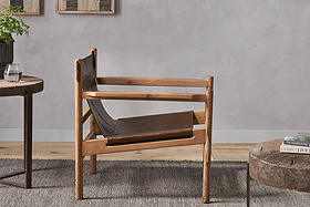 The Dalibar Leather Lounger - Tan by nkuku, featuring a wood frame and leather sling seat, sits on a textured gray rug with a round side table and low coffee table, creating a modern minimal look against the light gray wall.