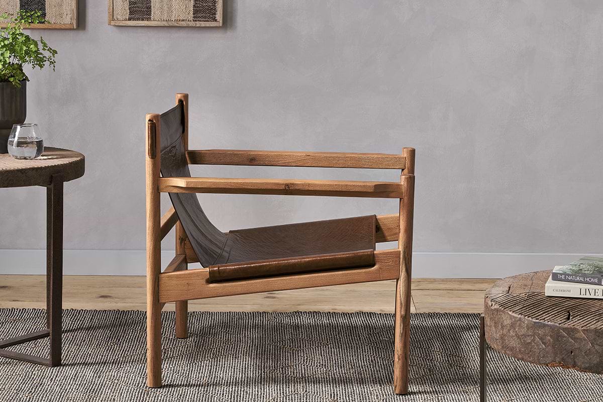 The Dalibar Leather Lounger - Tan by nkuku, featuring a wood frame and leather sling seat, sits on a textured gray rug with a round side table and low coffee table, creating a modern minimal look against the light gray wall.